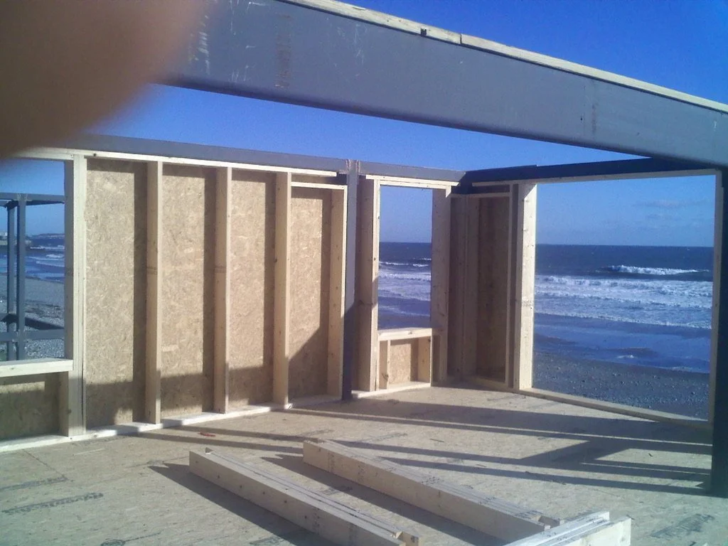 Wooden frame construction on a beachside, with the ocean and waves visible in the background, under a clear blue sky.