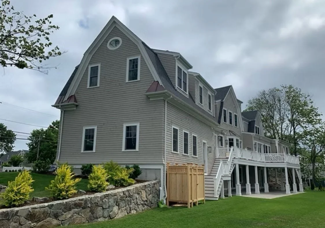 Large multi-story beige house with a stone foundation, white trim, and multiple windows, surrounded by a green lawn and landscaped bushes, under a cloudy sky.