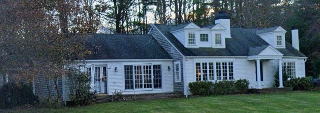 White two-story house with black roof, multiple large windows, and a small porch at the front. Surrounded by green lawn and tall trees.