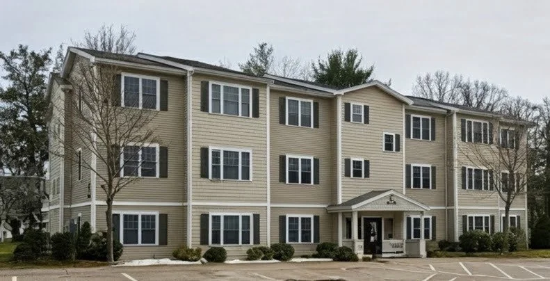 A three-story beige apartment building with multiple windows, a central entrance with a small porch, and parking spaces 