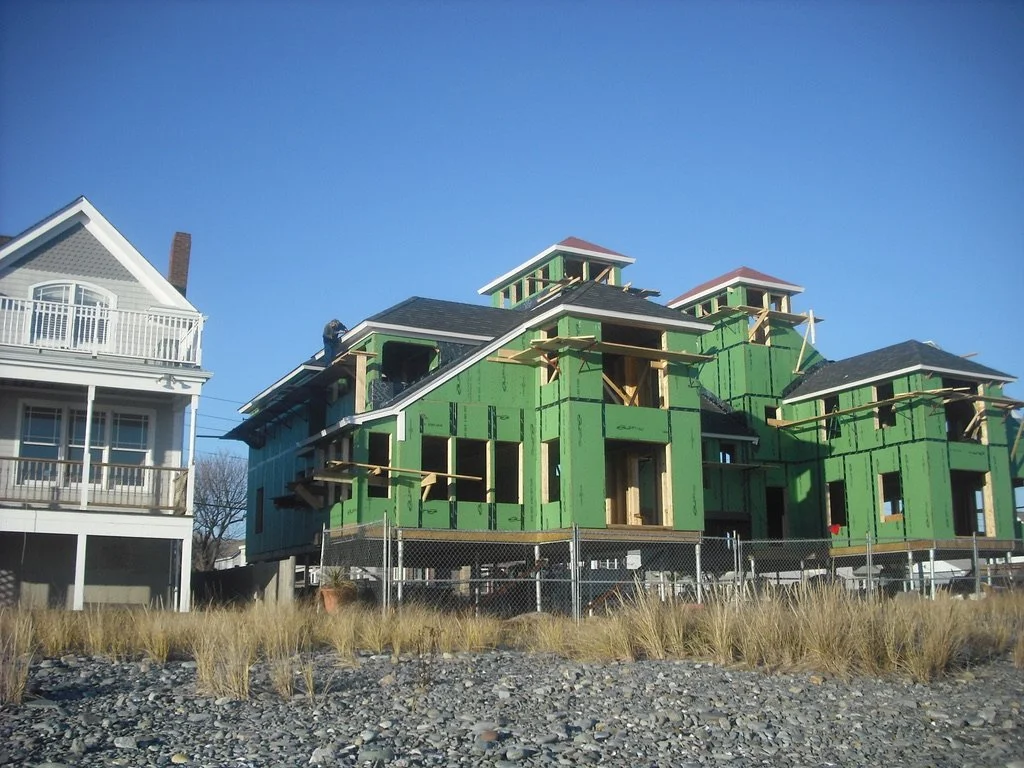 A house under construction with green sheathing, open window spaces, and no exterior siding, located next to a completed house with a porch and balcony, on a grassy and rocky area 