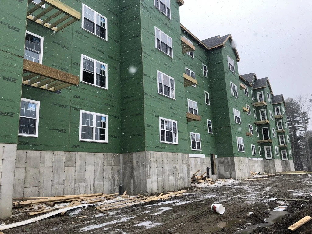 Multi-story residential building under construction with green exterior sheathing, white-framed windows, and a concrete foundation,
