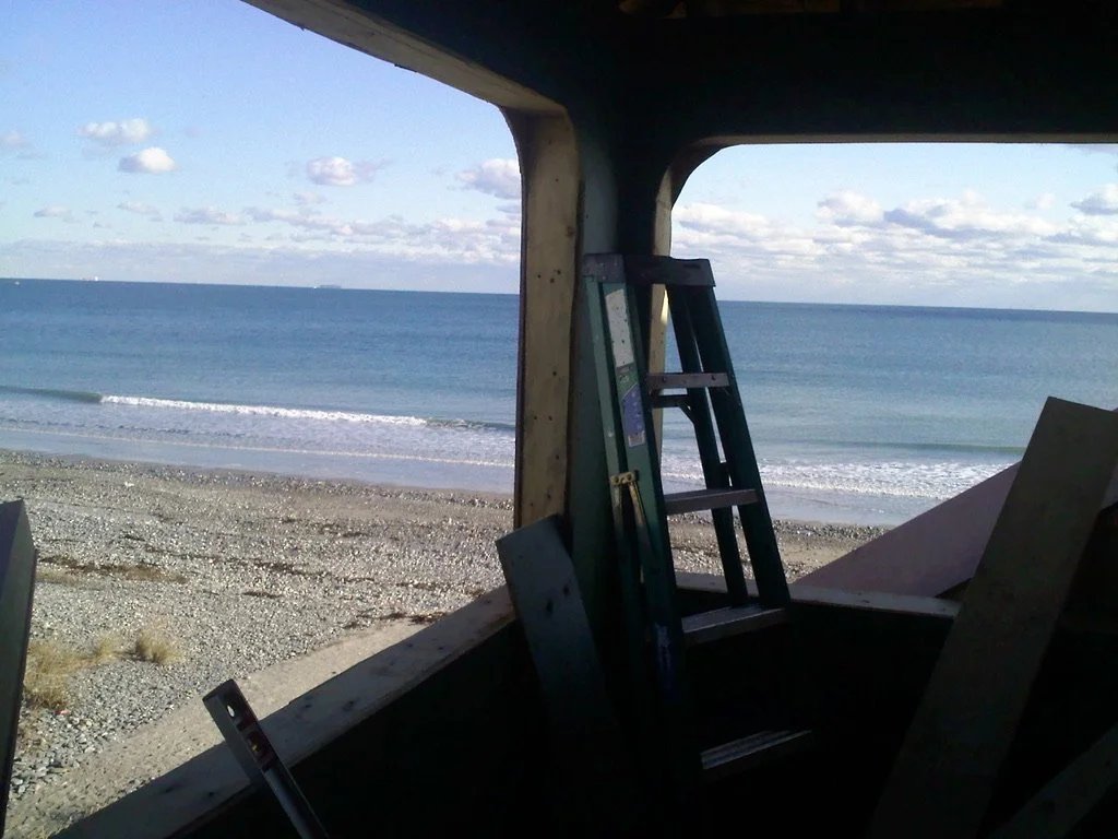 View through a beach hut window showing the ocean, sandy and pebbled shoreline, with a ladder leaning inside the hut.
