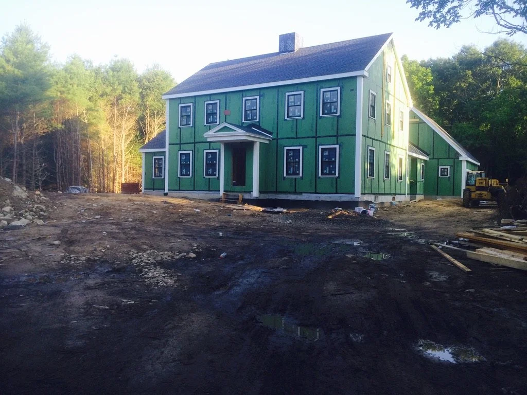 A two-story house under construction with green sheathing and black window trim, located in a wooded area, with construction materials and machinery around.