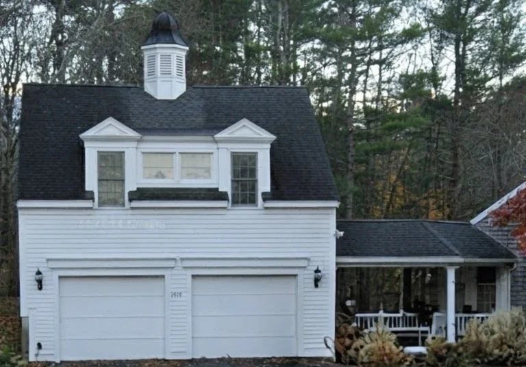 White garage with a small house attached, surrounded by trees, with a porch on the right side.