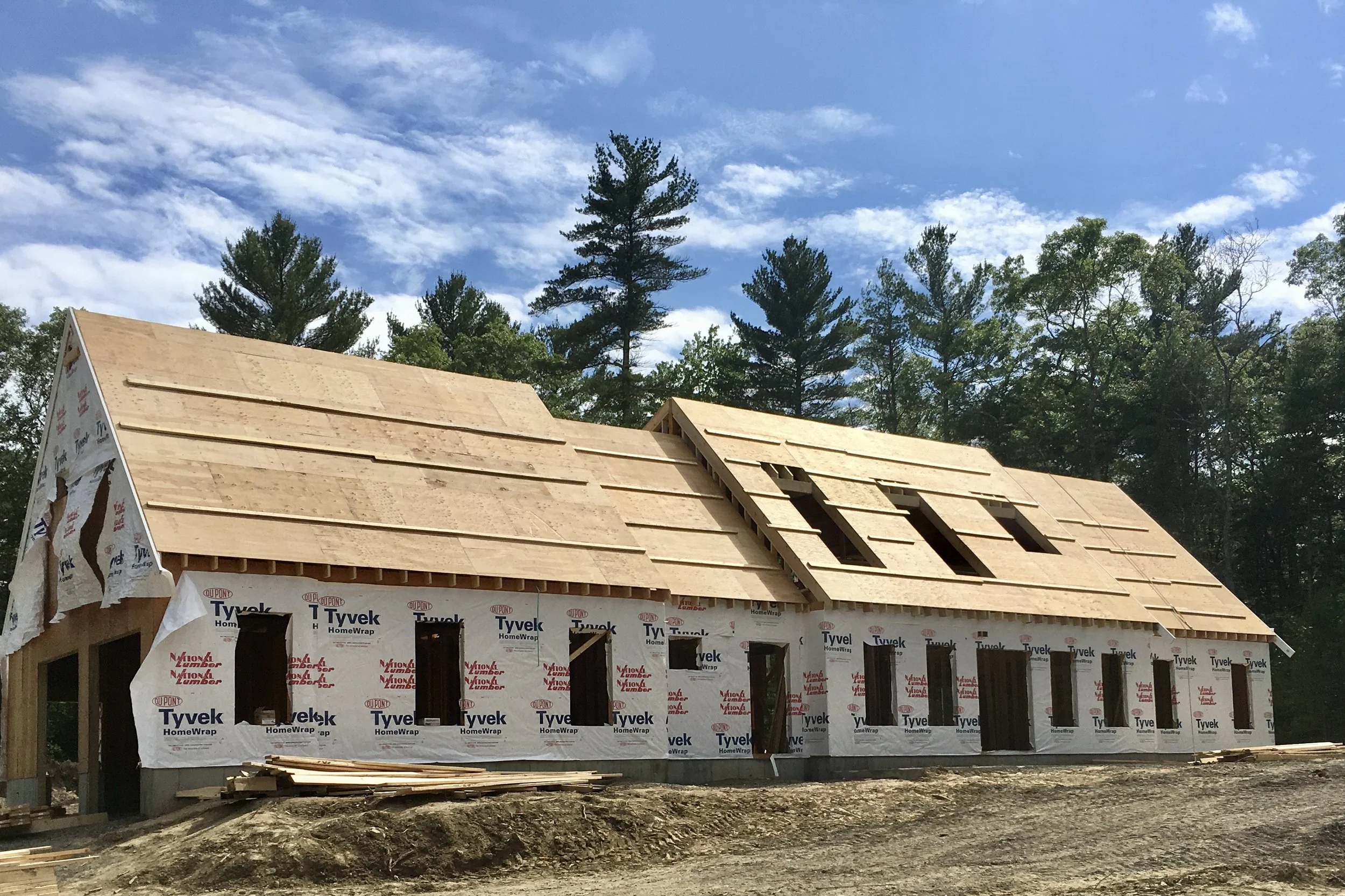 A house under construction with a wooden roof and wrapped exterior, set against a background of trees and a blue sky with clouds.