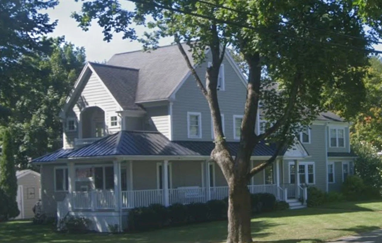 A large two-story house with gray siding and white trim, surrounded by trees and a well-maintained lawn, featuring a spacious covered porch and multiple windows.