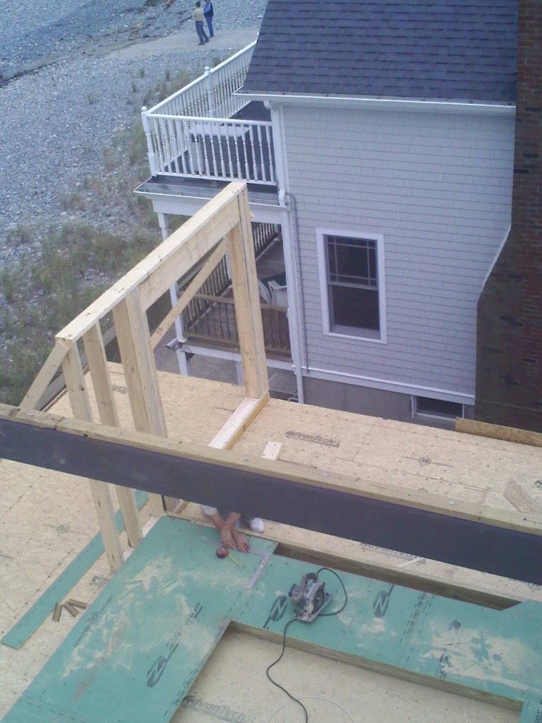 Construction site on a house deck with wooden framing, power tools, and a partially built railing, overlooking neighboring houses and a gravel area.