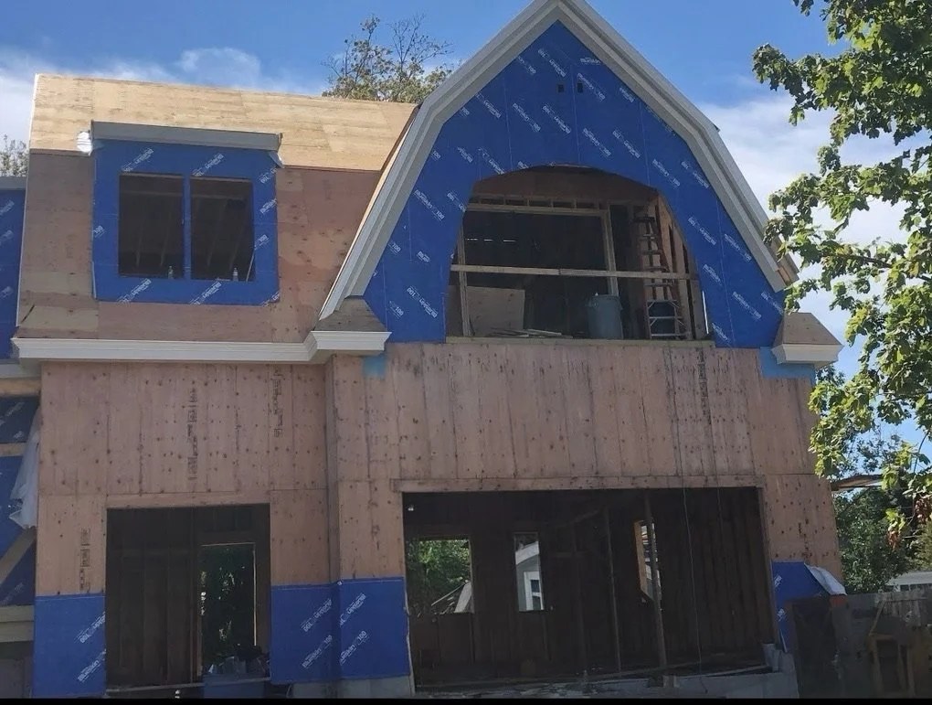 A house under construction with wooden framing, blue protective wrap, partially finished roof, and opening for windows and a balcony, with trees and a blue sky in the background.