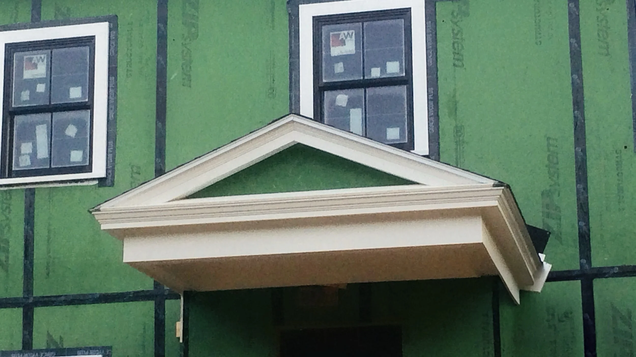 Close-up of a house under construction showing green sheathing, windows, and a decorative white gable roof above the entrance.