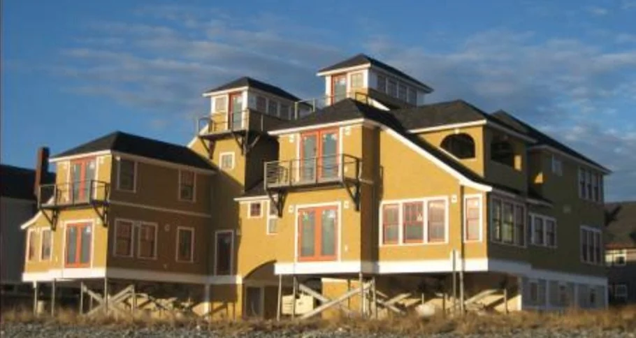 A large, colorful multi-level house with yellow and brown exterior, multiple windows, balconies, and a stairway, set against a blue sky.