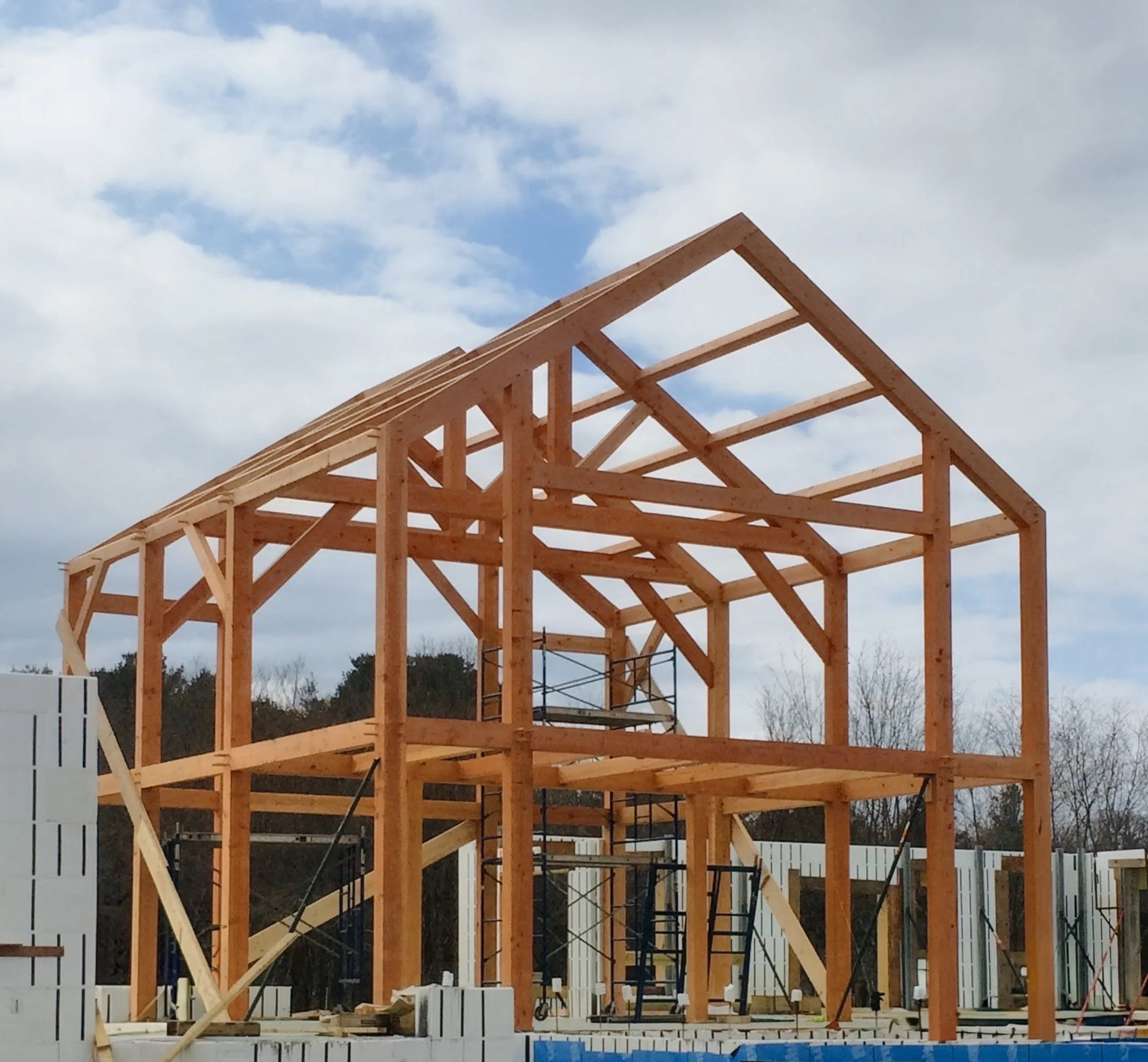 Wooden house frame under construction with scaffolding and construction materials around, blue sky with clouds in the background.