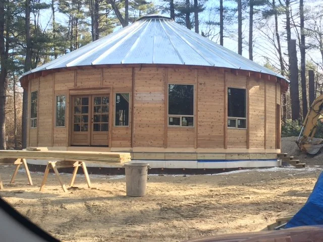 Circular wooden structure under construction with a metal roof, surrounded by trees, with construction tools and materials nearby.