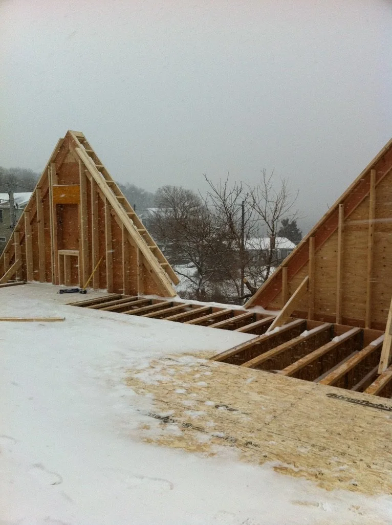 Construction site with wooden framework for a roof in snowy weather, neighboring houses and leafless trees in the background.