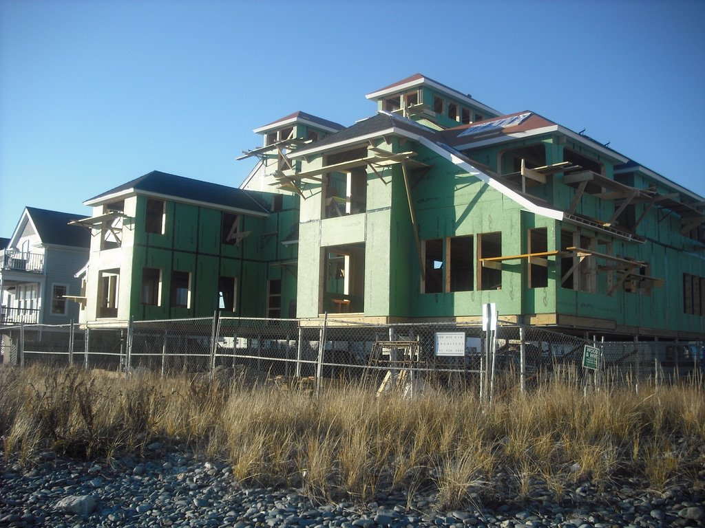 Under-construction multi-story house with green sheathing, wooden support structures, and sloped roofs, fenced off with construction site signage, located near grassy and rocky terrain.