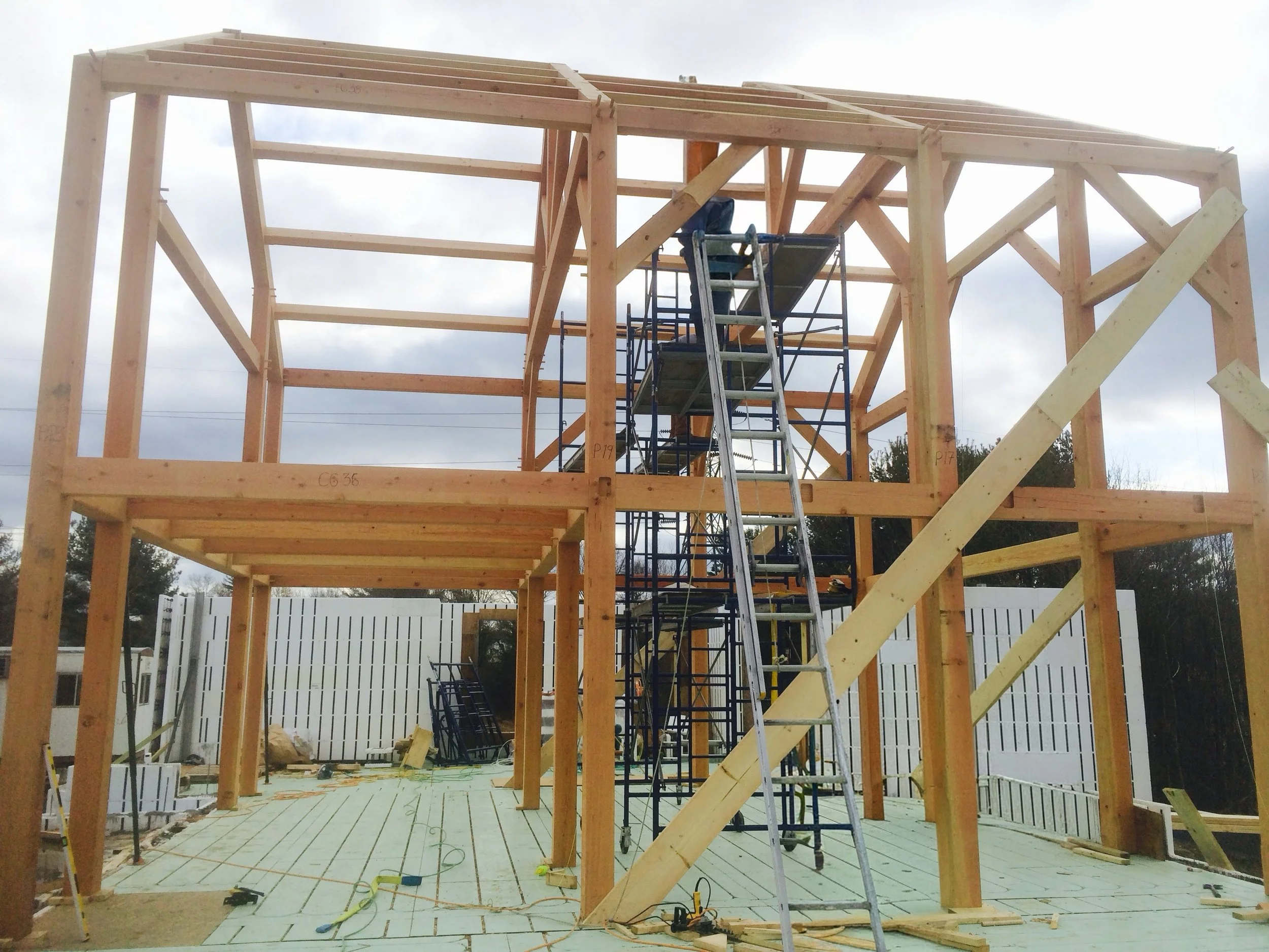 Wooden house framing under construction with scaffolding and ladder, construction materials scattered on the floor, and white fencing in the background.
