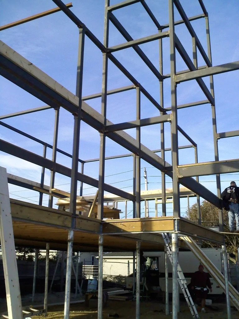 Construction site with metal framing for a building, wooden platforms, and workers assembling the structure under a blue sky.