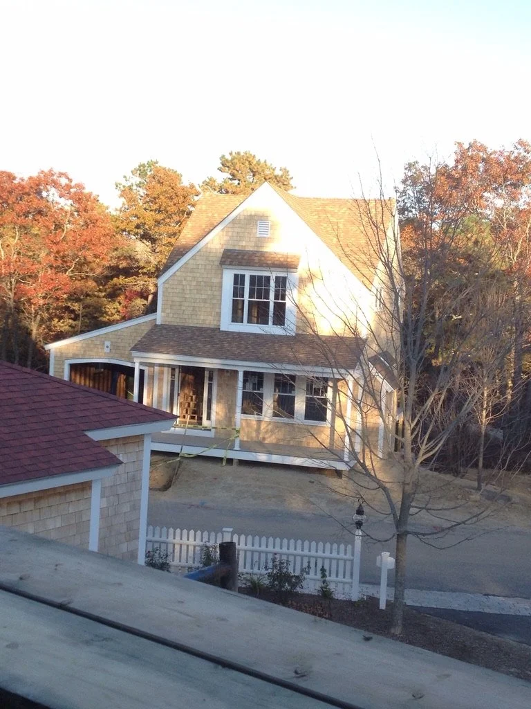 A two-story house under construction with beige siding, multiple windows, and a gabled roof, located in a suburban neighborhood