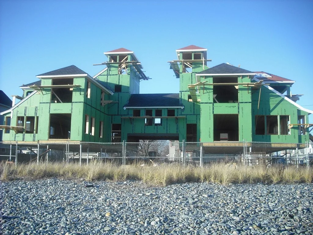 Under-construction house with green sheathing, multiple levels, and small towers, located behind a chain-link fence on a rocky and grassy area.