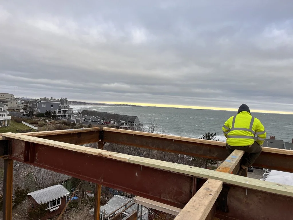 A construction worker in a bright yellow safety jacket and dark hood sits on a steel beam frame of a building under construction, overlooking a coastal neighborhood with houses and the ocean in the distance during a cloudy day.