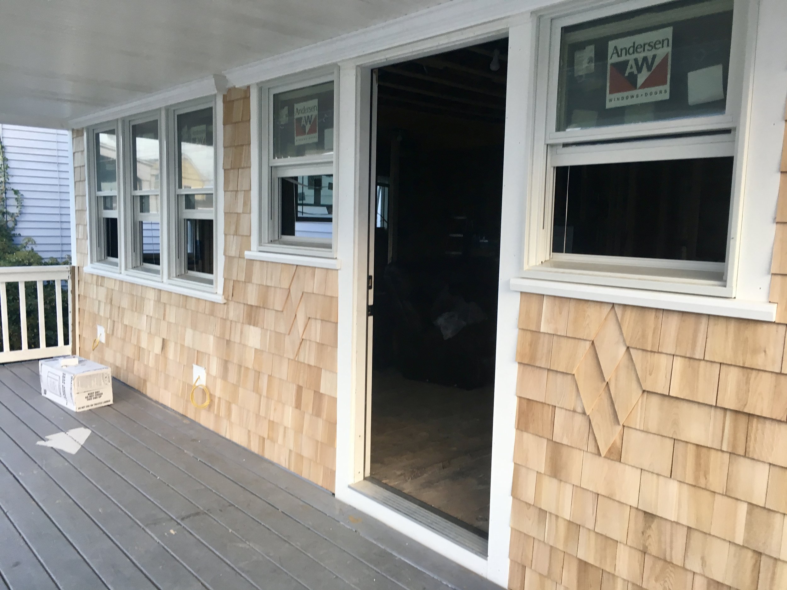 Exterior of a house under construction with unfinished window frames and shingle siding, a sliding glass door open to the interior, and a deck 