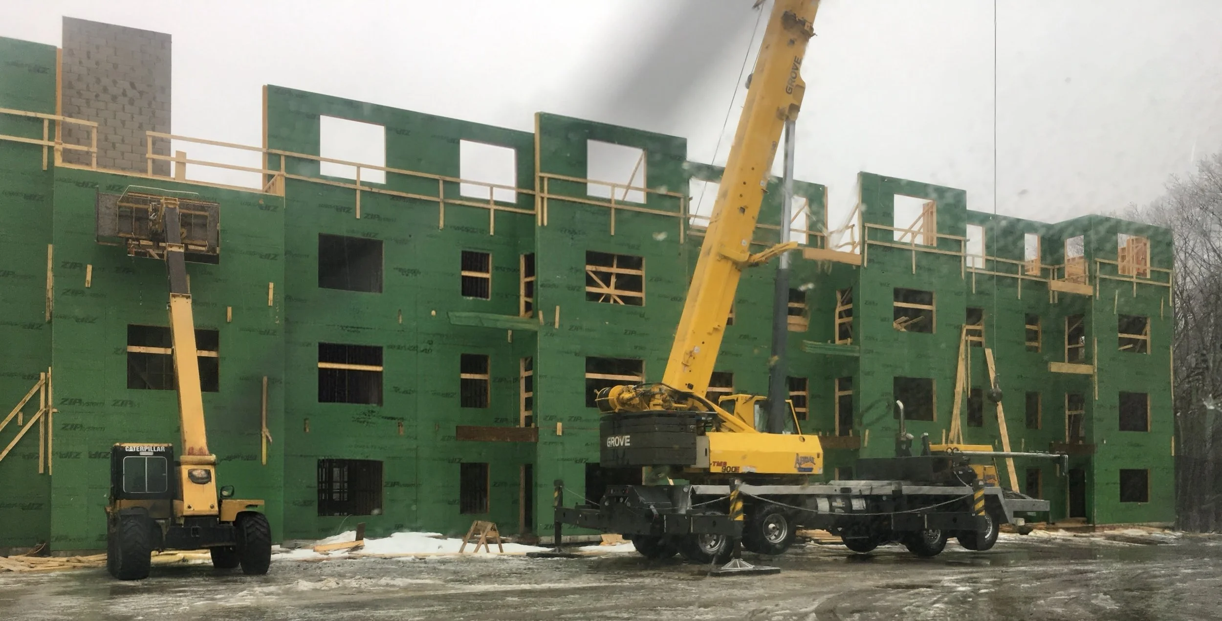 Construction site with a partially built multi-story building covered in green sheathing, with construction cranes and equipment in the foreground