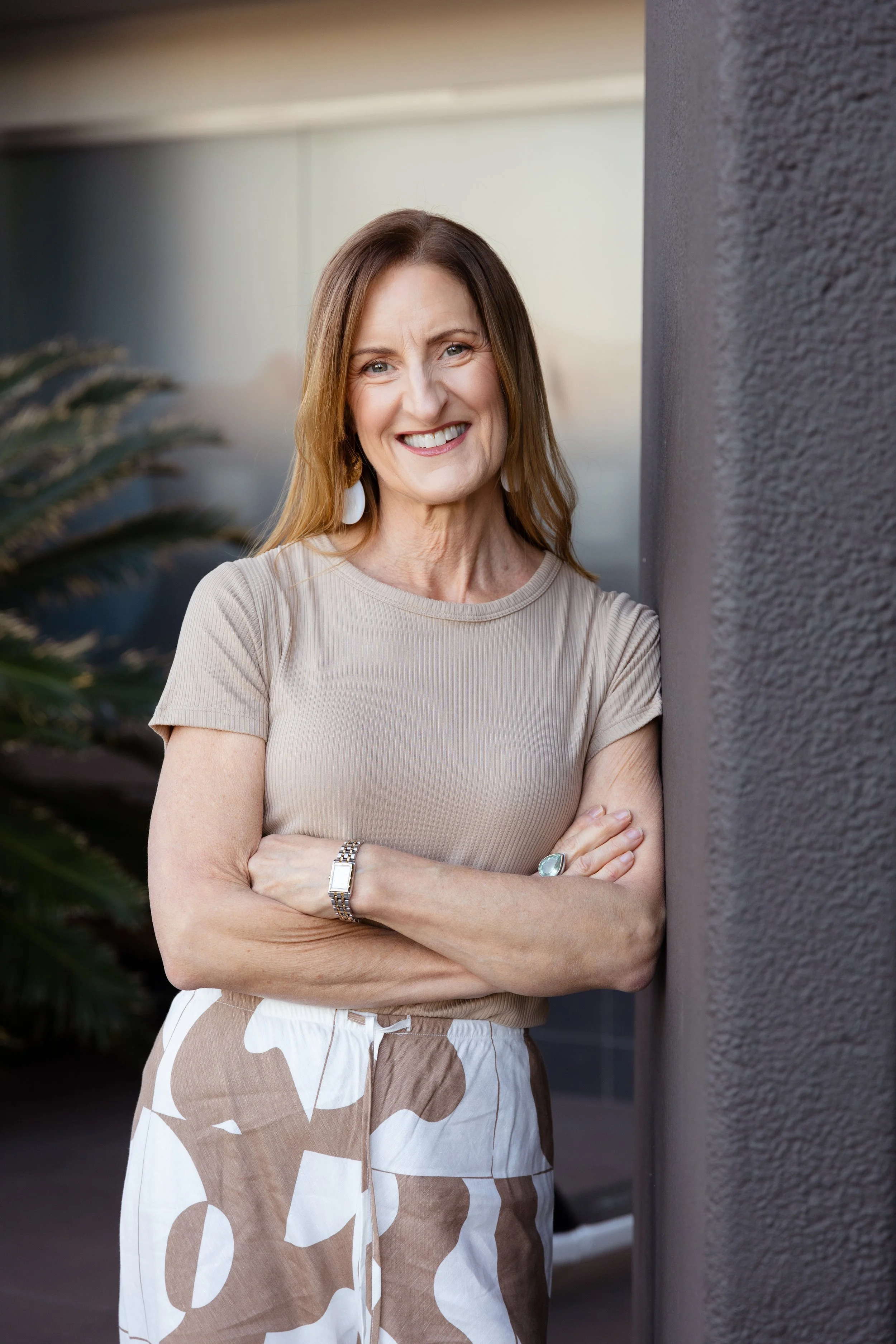 A smiling woman with shoulder-length brown hair standing outdoors, leaning against a textured wall with her arms crossed, wearing a beige ribbed t-shirt and patterned beige and white pants.