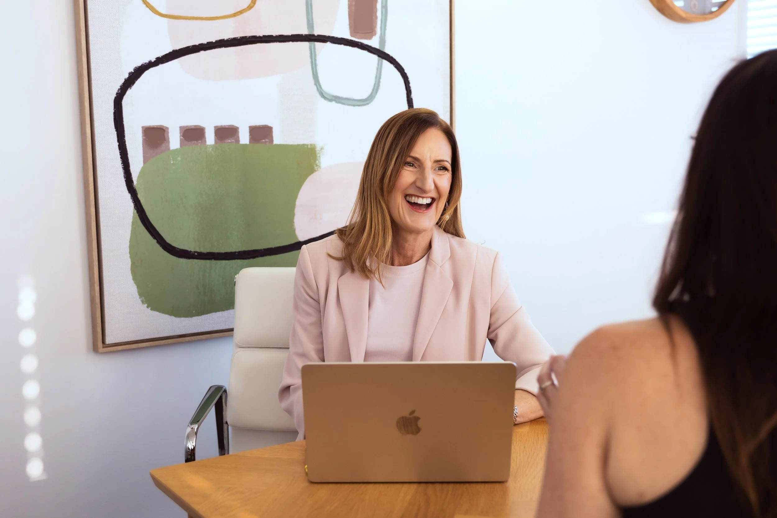 Two women having a conversation in an office, with one smiling and the other partially visible, sitting at a wooden desk with a silver MacBook.