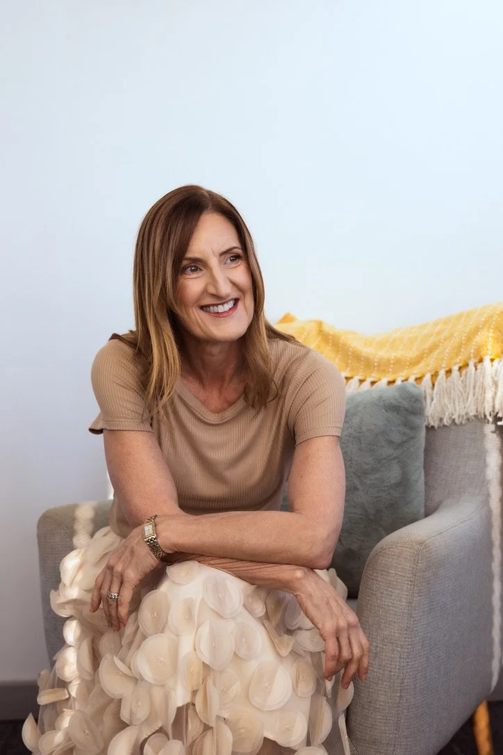 A woman in a beige top and cream-colored skirt sitting on a gray couch, smiling in a bright room with a light blue wall and a yellow throw blanket in the background.