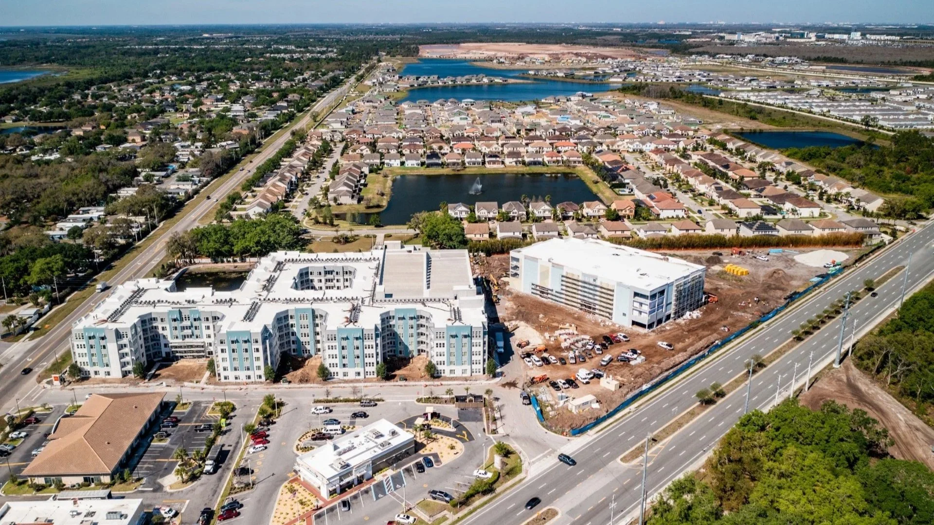 Aerial view of a residential area with ongoing construction, surrounded by lakes and a highway.