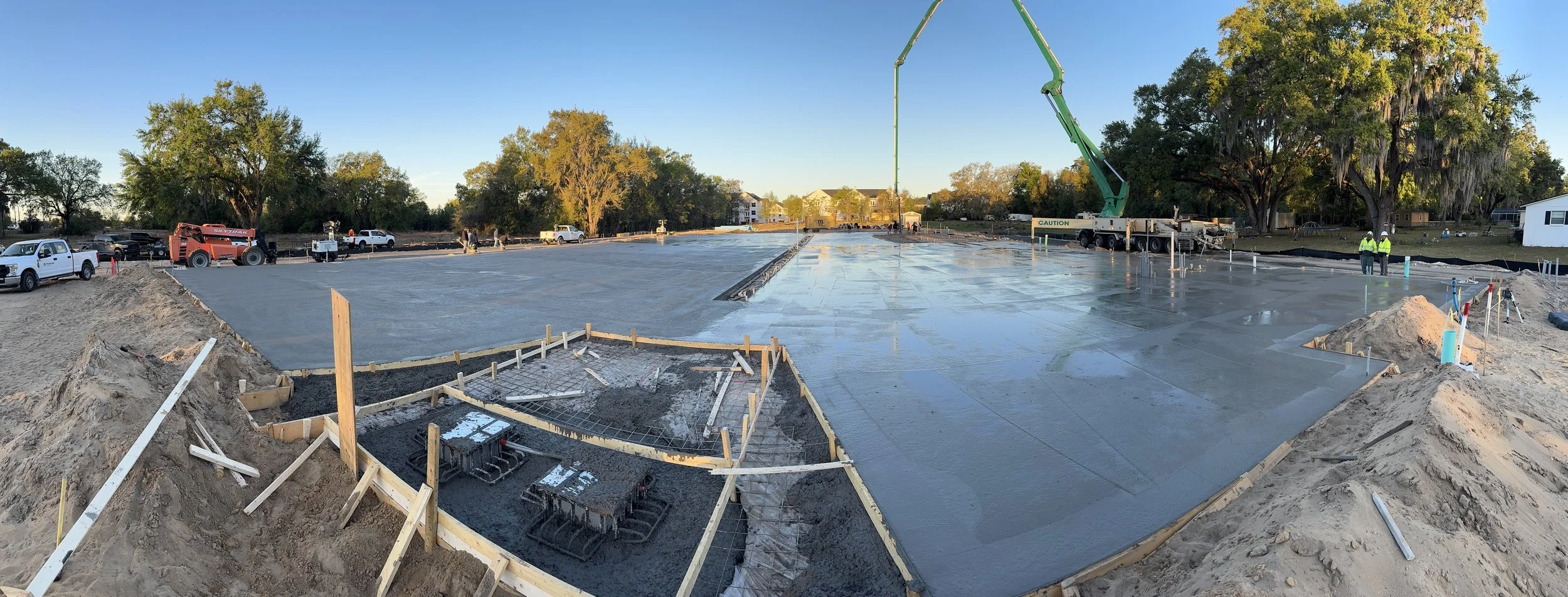 Construction site with freshly poured concrete slab for a building foundation, surrounded by construction equipment, trucks, and workers in safety vests and helmets, with trees and houses in the background.