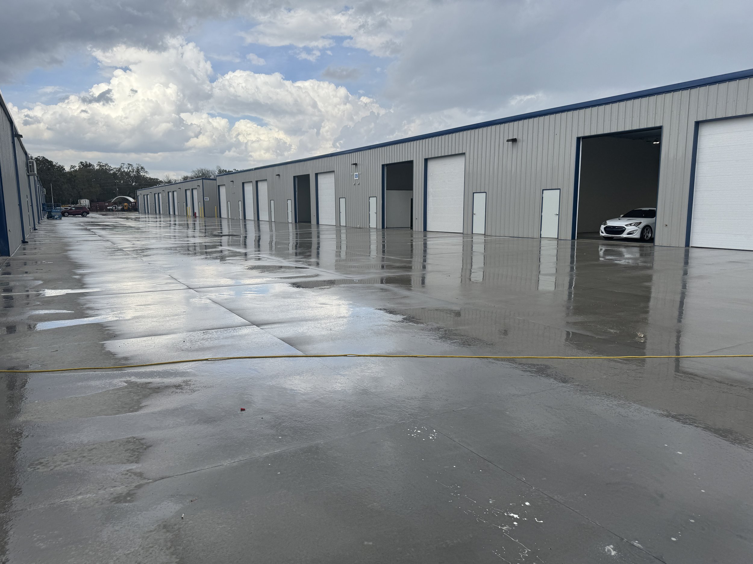 A row of industrial storage units with open garage doors, wet concrete ground reflecting the sky and buildings, and a white car visible inside one of the units on the right.
