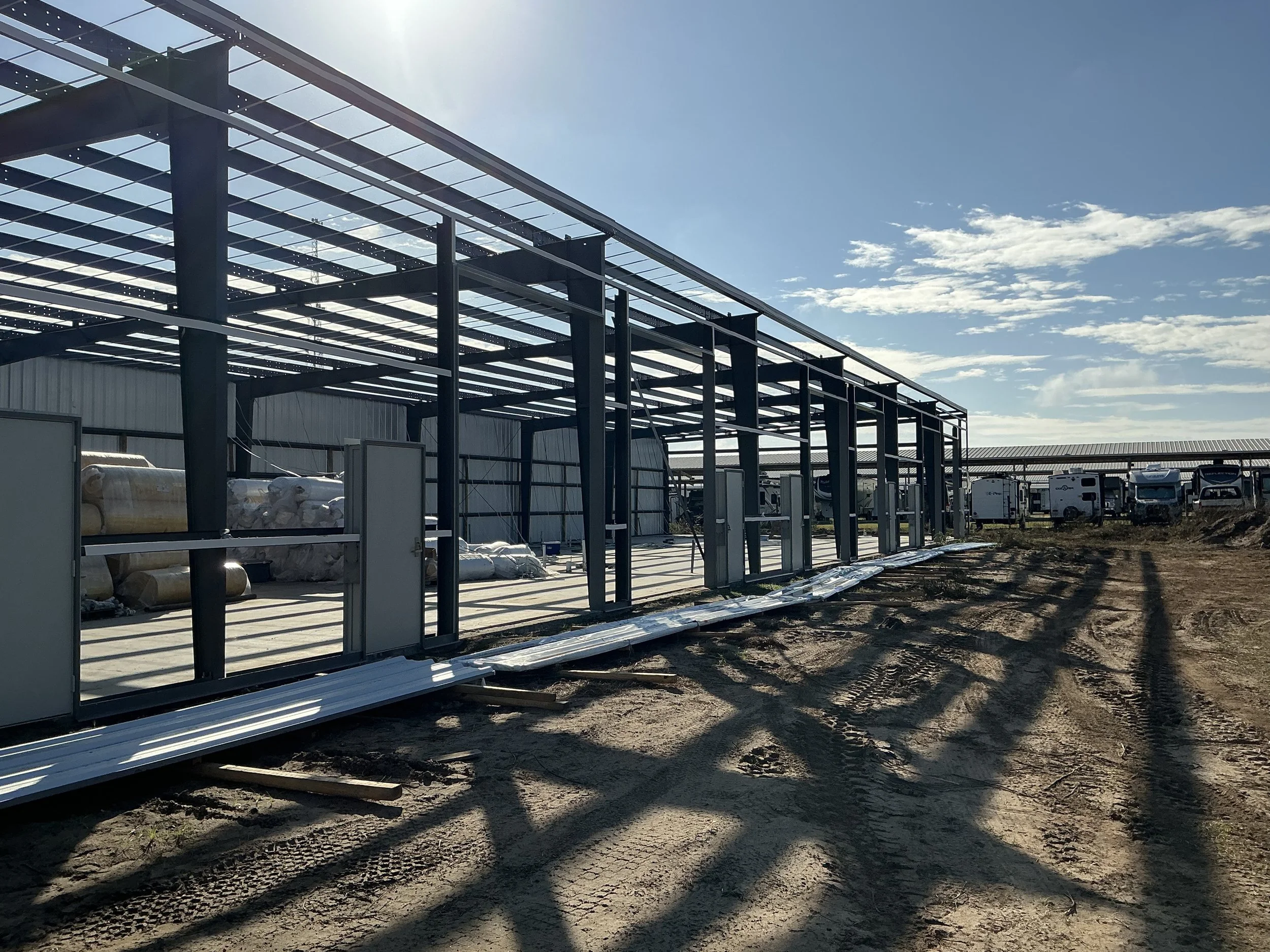 Construction site with steel framework for a building, dirt ground with tire marks, and parked RVs in the background under a partly cloudy sky.