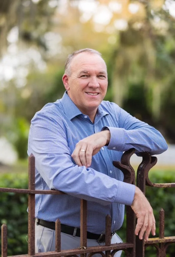 A man with light skin, short gray hair, wearing a blue dress shirt, leaning on a rusty metal gate in a park or garden with trees and greenery in the background.