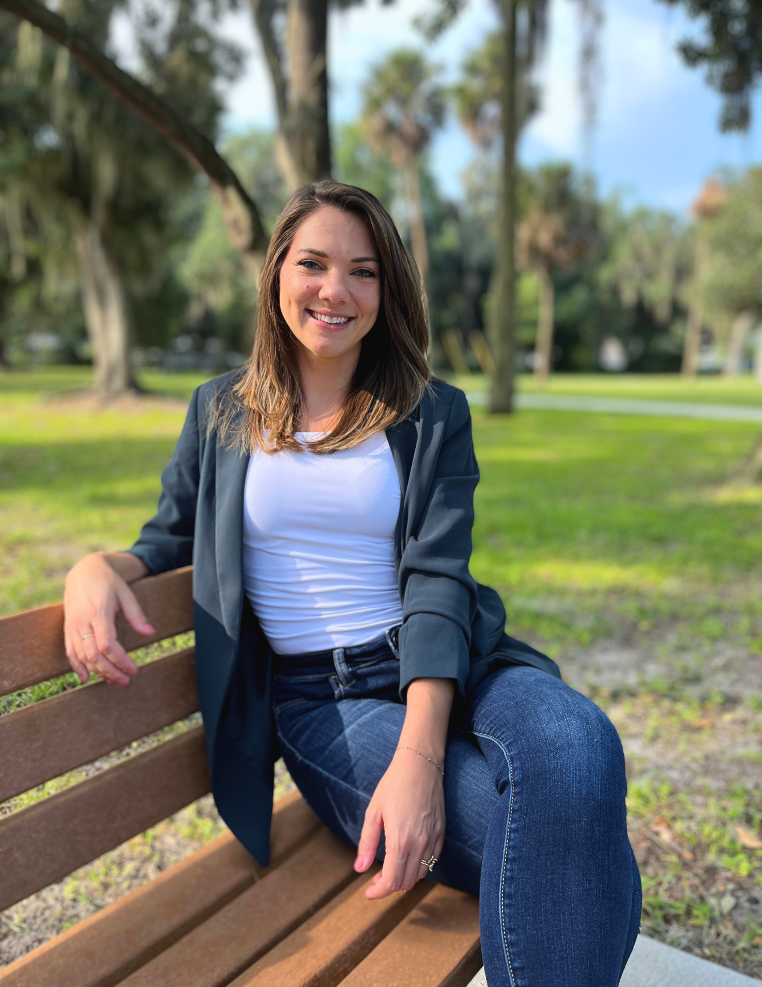 A young woman with brown hair, smiling, sitting on a wooden park bench in a lush green park during daytime.