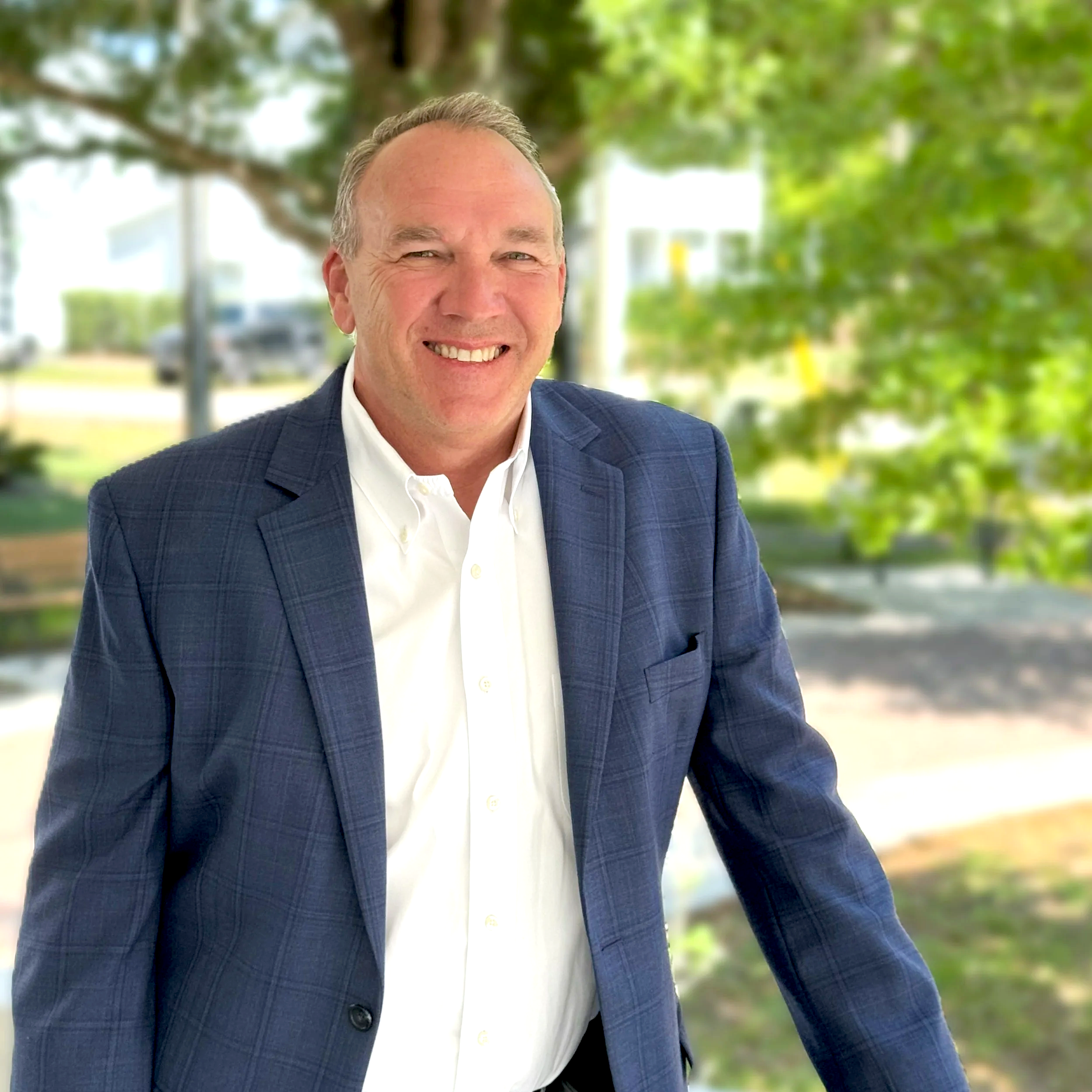 Smiling man in a suit outdoors with trees and buildings in the background.