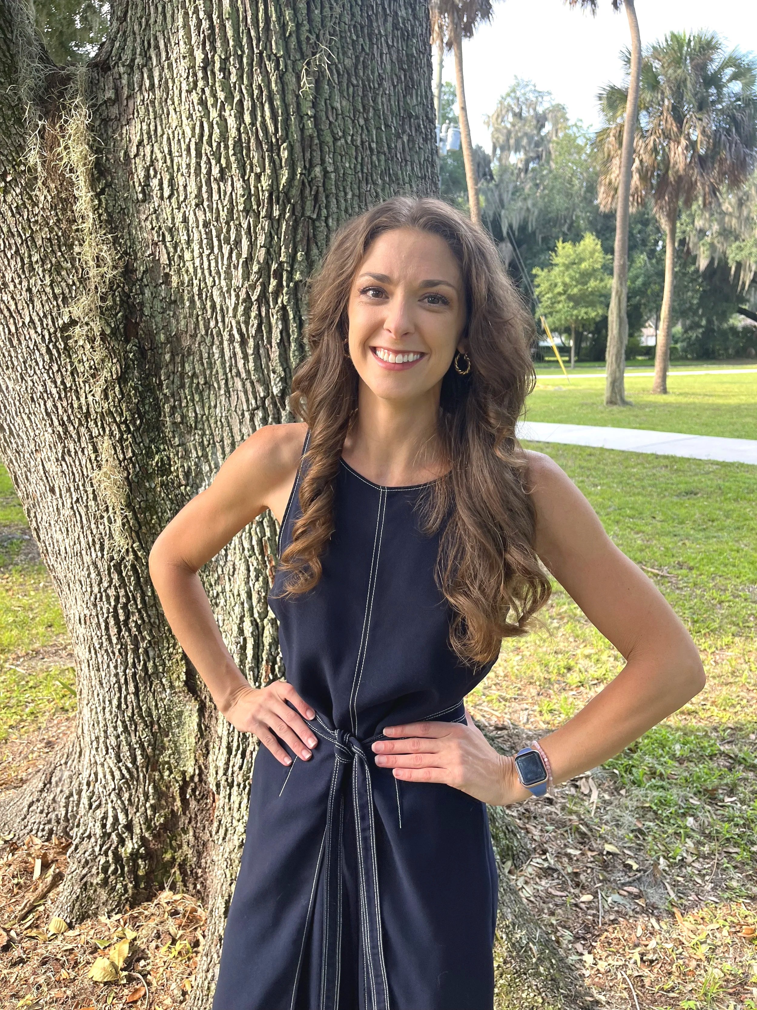 A woman with long, wavy brown hair in a sleeveless navy dress with a waist tie, smiling in front of a large tree in a park with grass, trees, and a sidewalk.