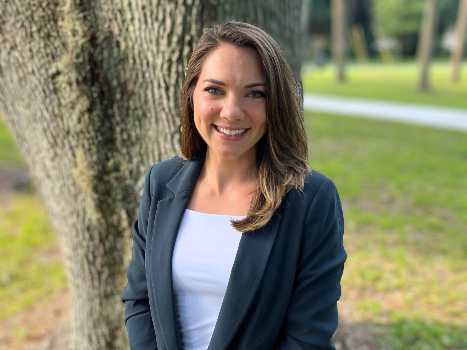 A smiling woman with long brown hair, wearing a blue blazer over a white top, standing outdoors near a large tree in a park.