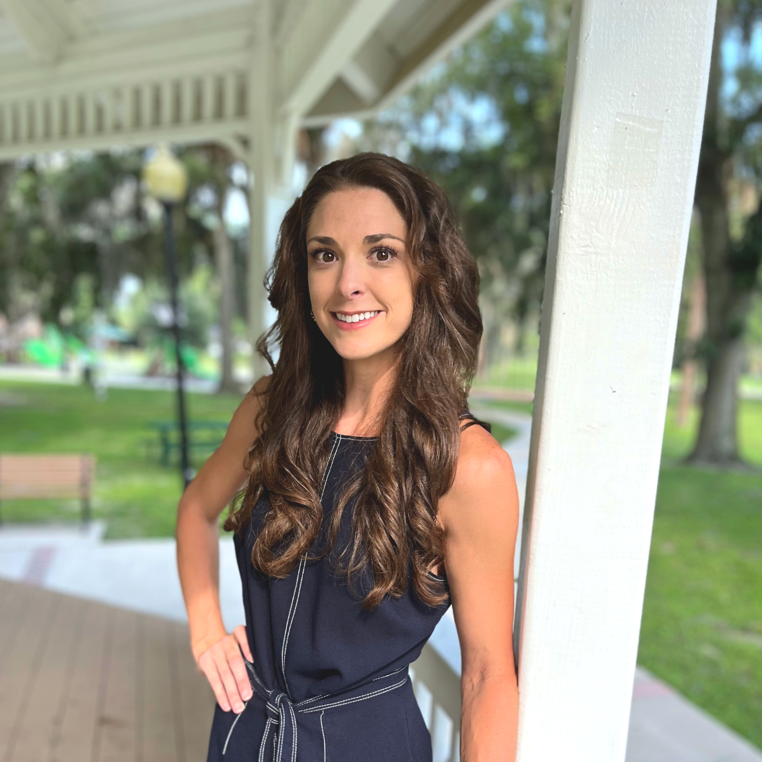 A young woman with long wavy brown hair, smiling, standing outside near a white wooden structure in a park with trees and benches in the background.