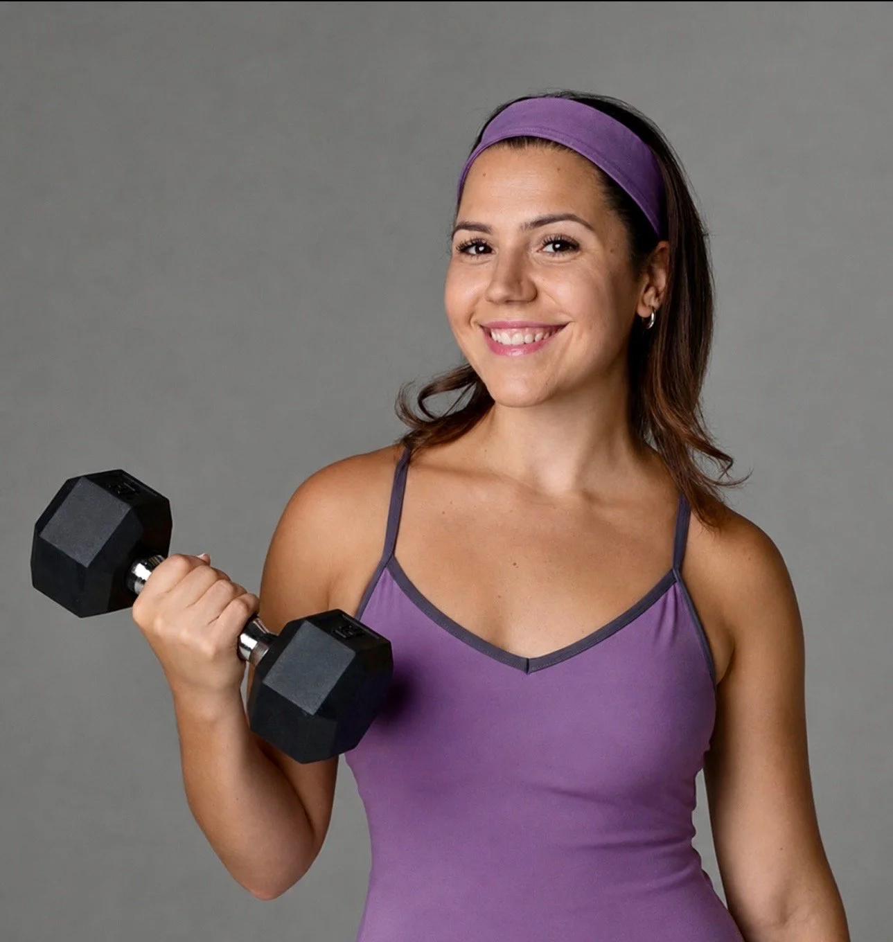 A young woman with a purple headband and matching tank top holding a black dumbbell and smiling against a gray background.