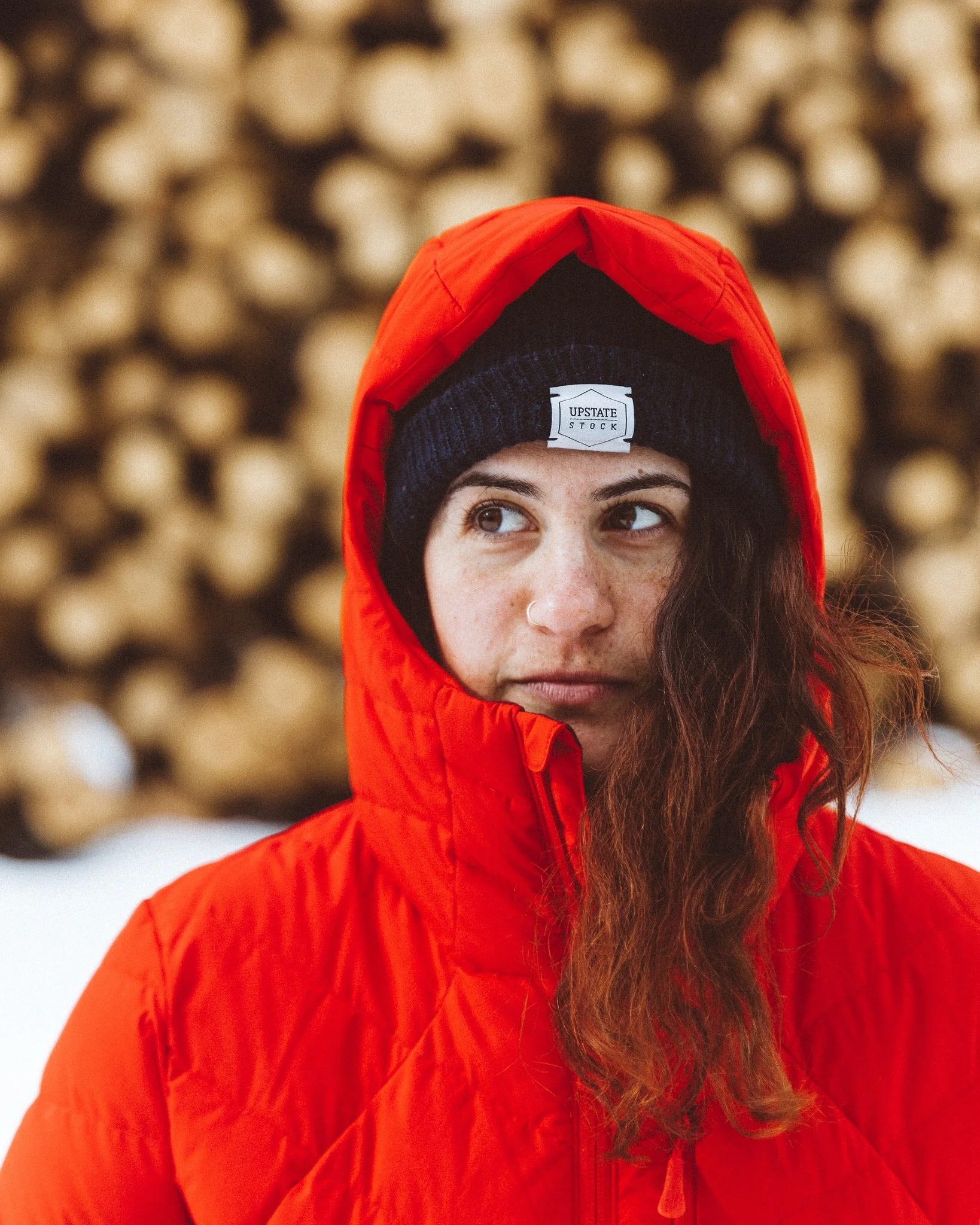 A person wearing a red winter jacket and a black beanie with a white label, standing outdoors with a blurred background of stacked logs.