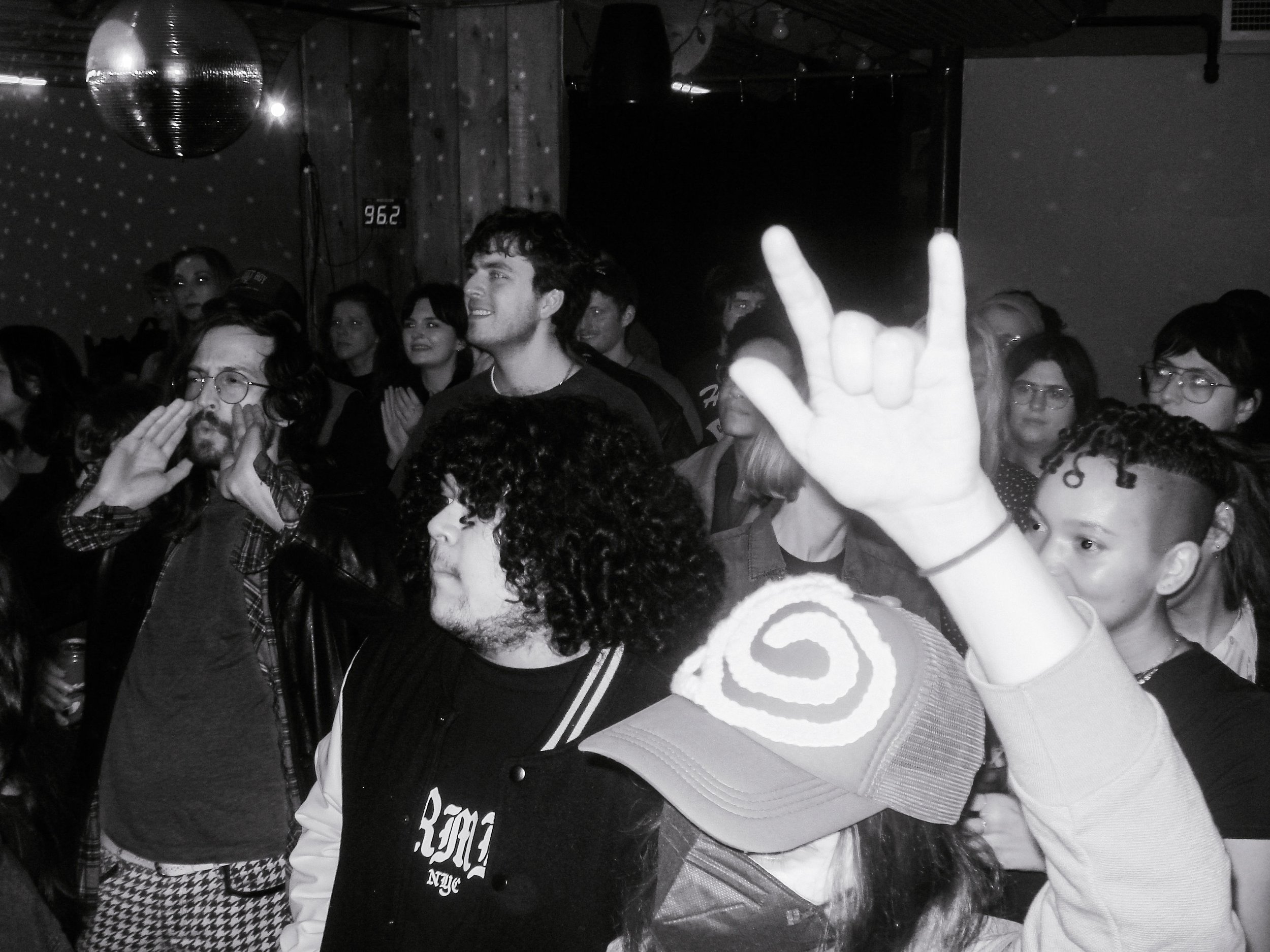 Stupid Lucky Live at Cassette in Ridgewood Queens, NYC - Photo by Beth Crane on Digital Camera. A black and white shot of the crowd, cheering and making the "rock" hand signal.