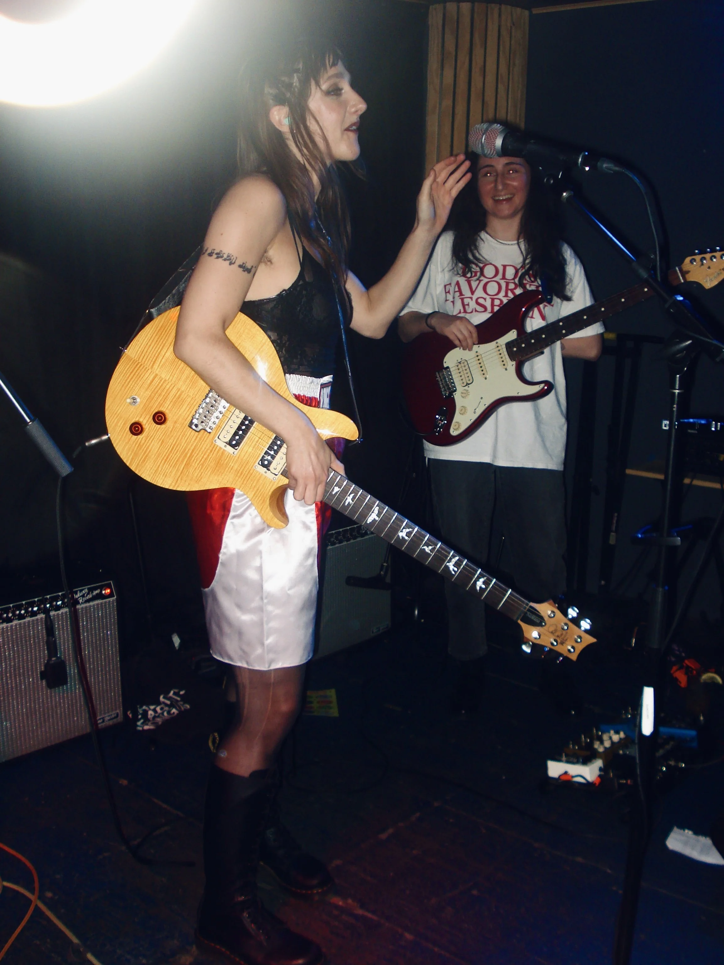 Stupid Lucky Live at Cassette in Ridgewood Queens, NYC - Photo by Beth Crane on Digital Camera. Taylor Pearlstein stands at the mic with her yellow guitar, and Fran stands in the background with her guitar.