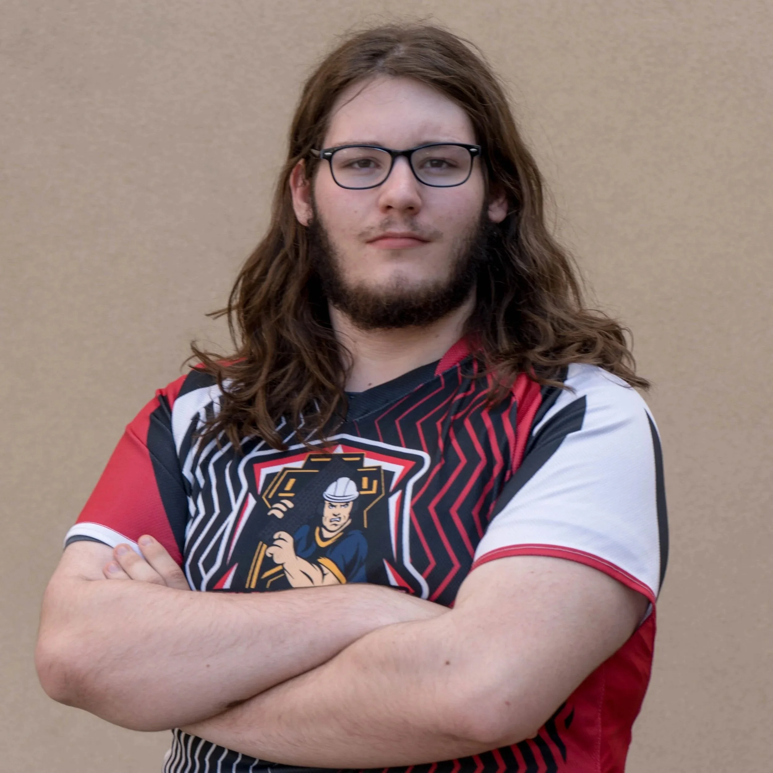A person with long brown hair, glasses, and a beard, wearing a sports jersey displaying a cartoon knight, crossing their arms in front of a plain beige wall.