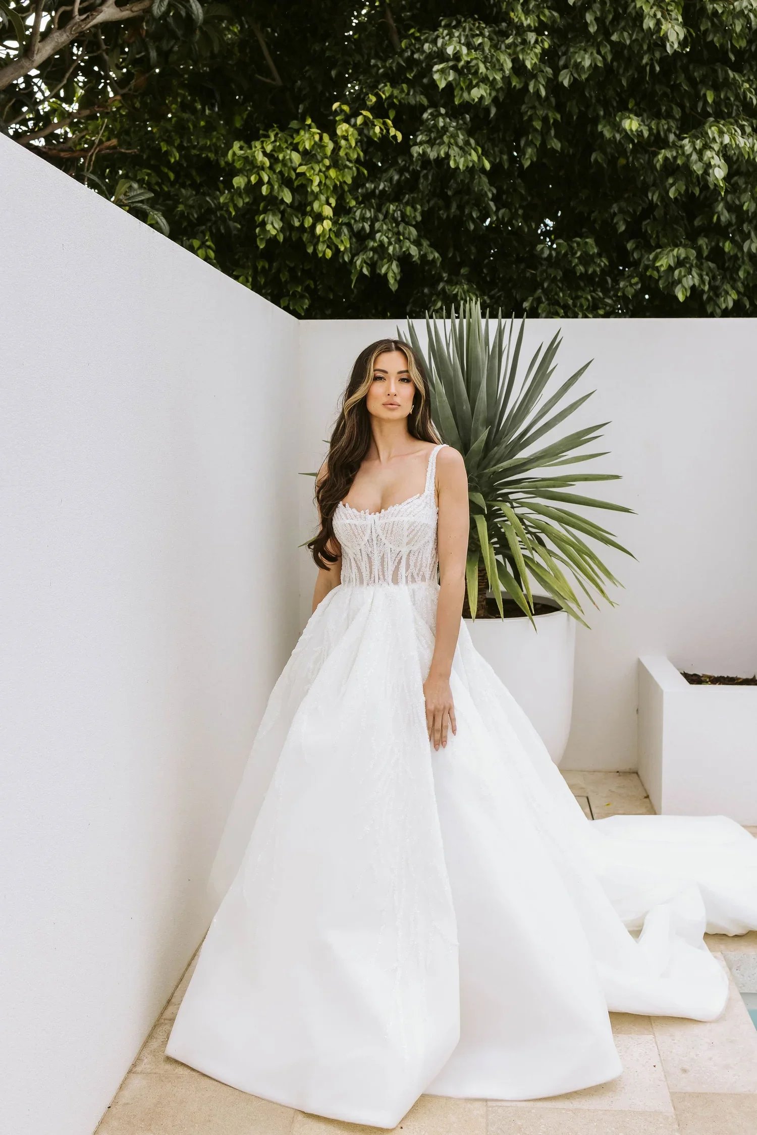 A woman in a white wedding dress stands next to a large green plant, with a white wall and greenery in the background.
