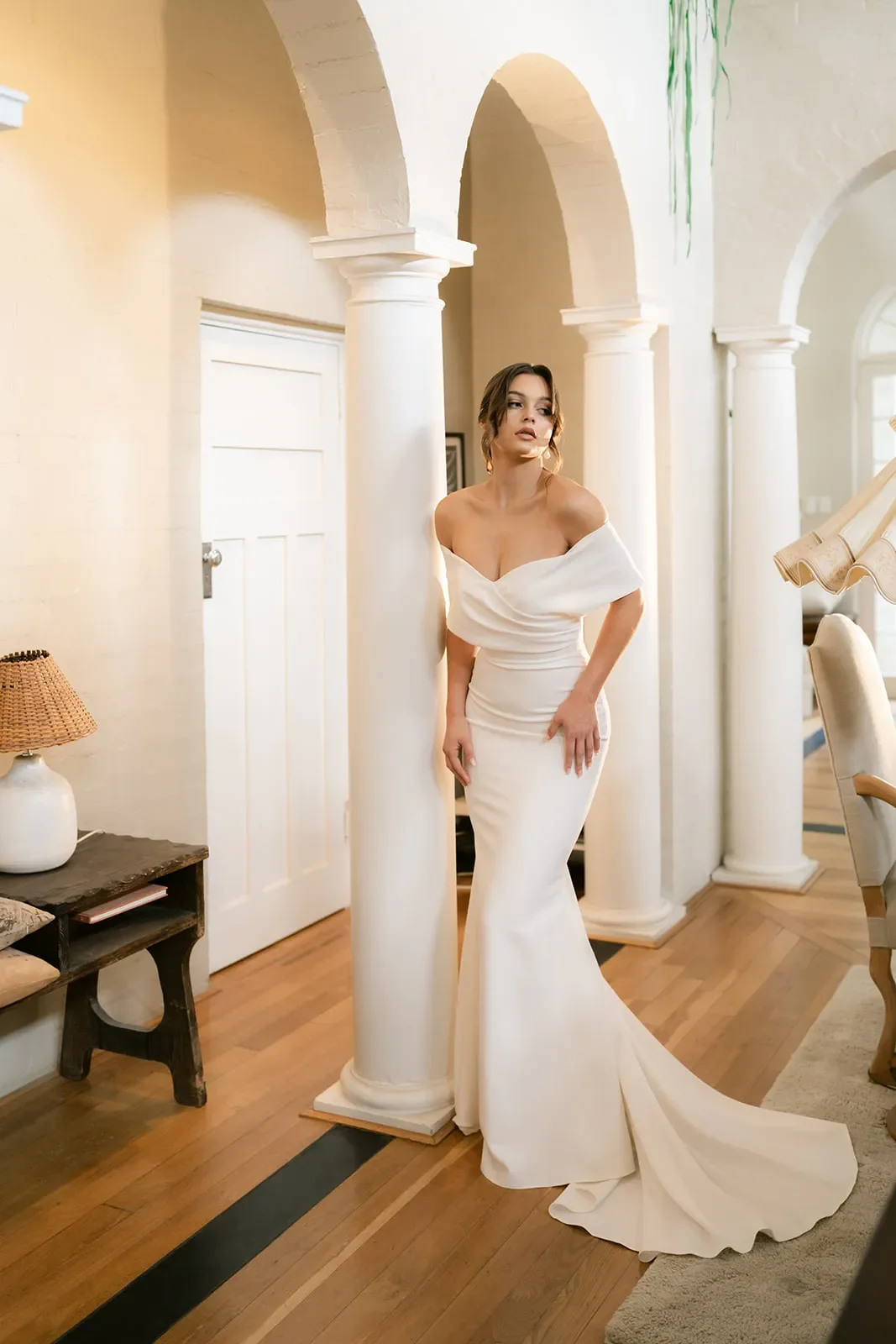 A woman in a white off-shoulder wedding gown standing next to a white column in a well-lit room with wooden flooring and decor.