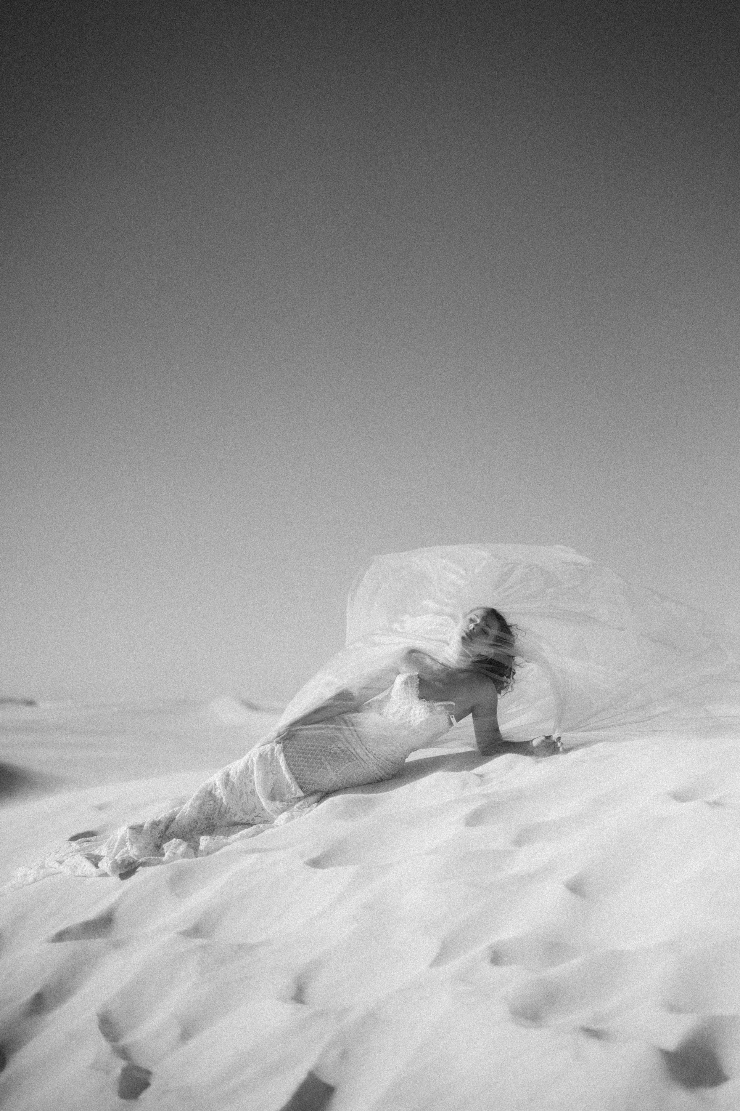 A person lying on the sand at a beach, wearing a light dress and holding a large parasol, with dunes in the background and a clear sky.