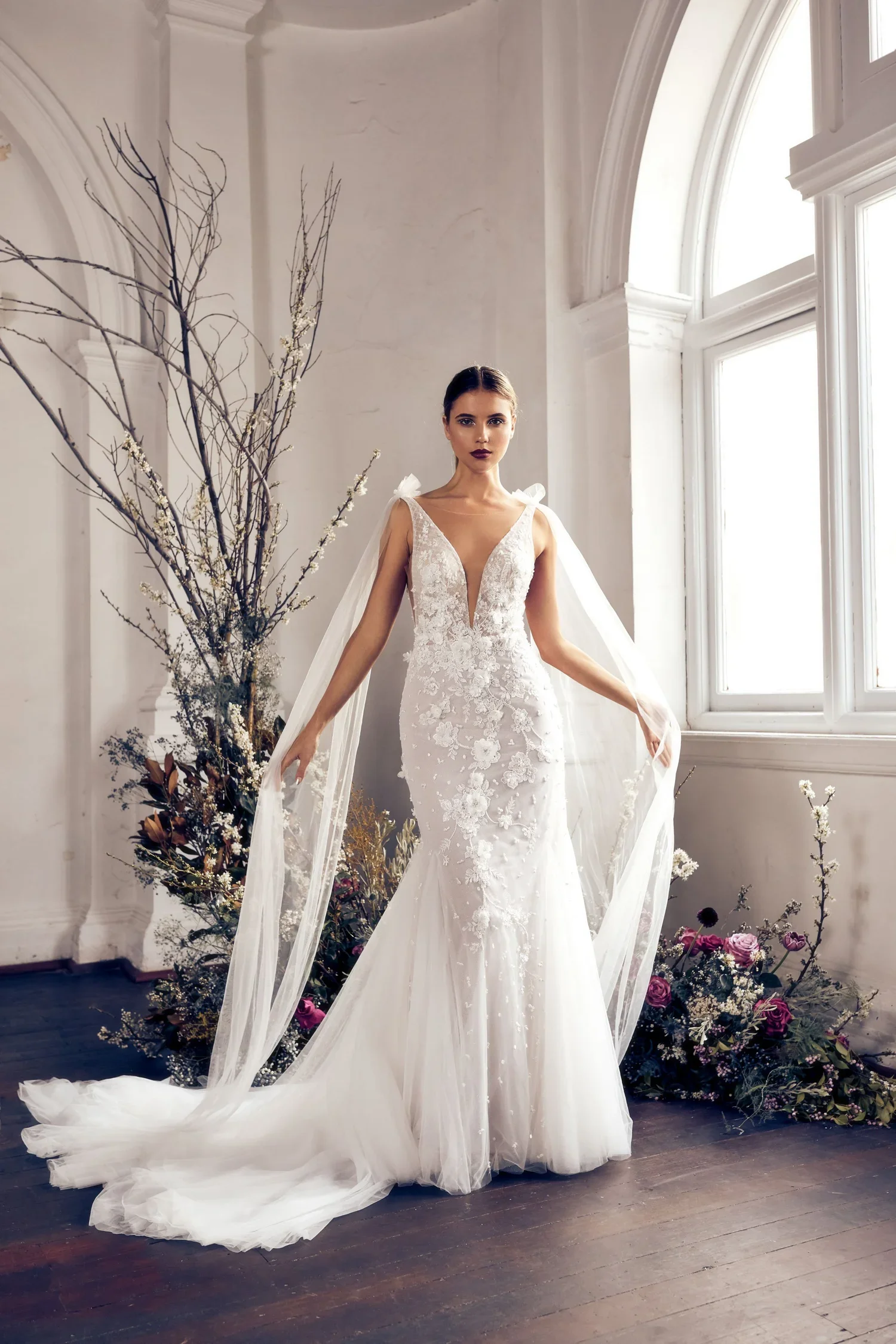 A woman in a white wedding gown with lace and floral details, standing indoors near large windows with white trim. She has dark hair styled back, dark lipstick, and is surrounded by floral arrangements with branches and roses.