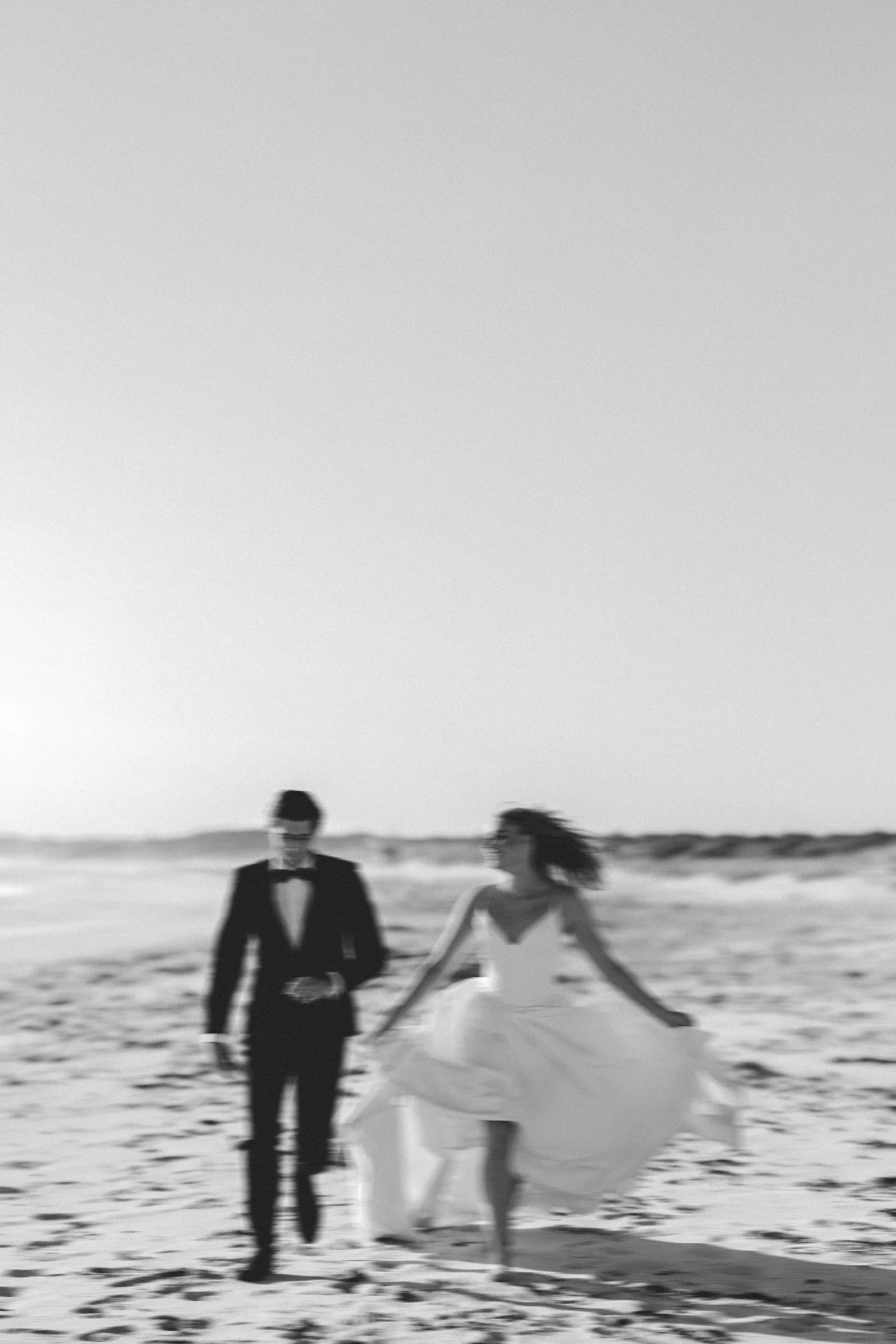 A black and white photograph of a couple on the beach, with the woman in a wedding dress holding a bouquet and the man dressed in a tuxedo.