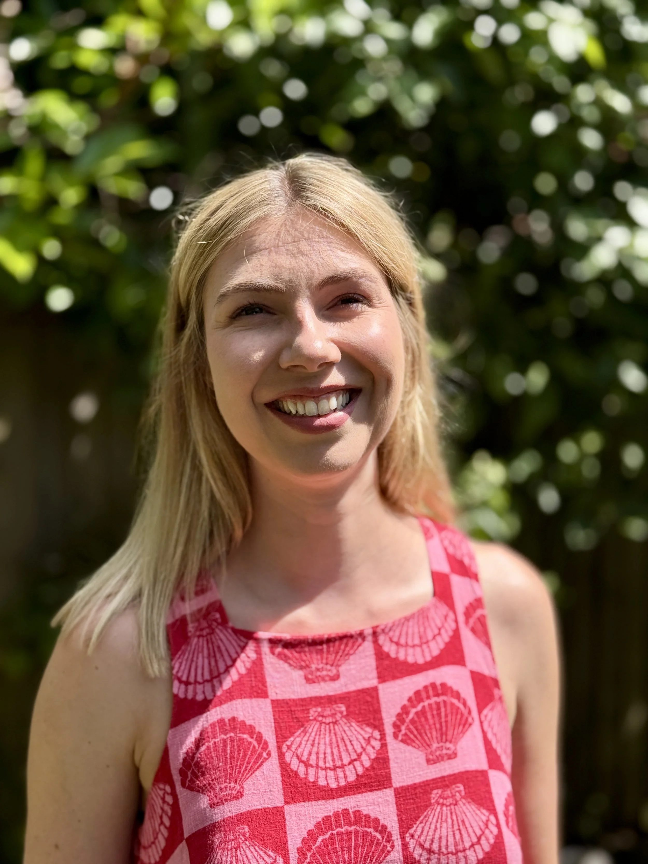 Young woman with long blonde hair smiling outdoors, wearing a sleeveless pink top with a seashell pattern, with green foliage in the background.
